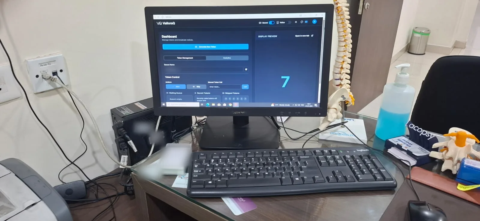 ValloraQ dashboard in dark mode on a desktop monitor in a doctor’s cabin, next to a spine model, with token 7 in the display preview