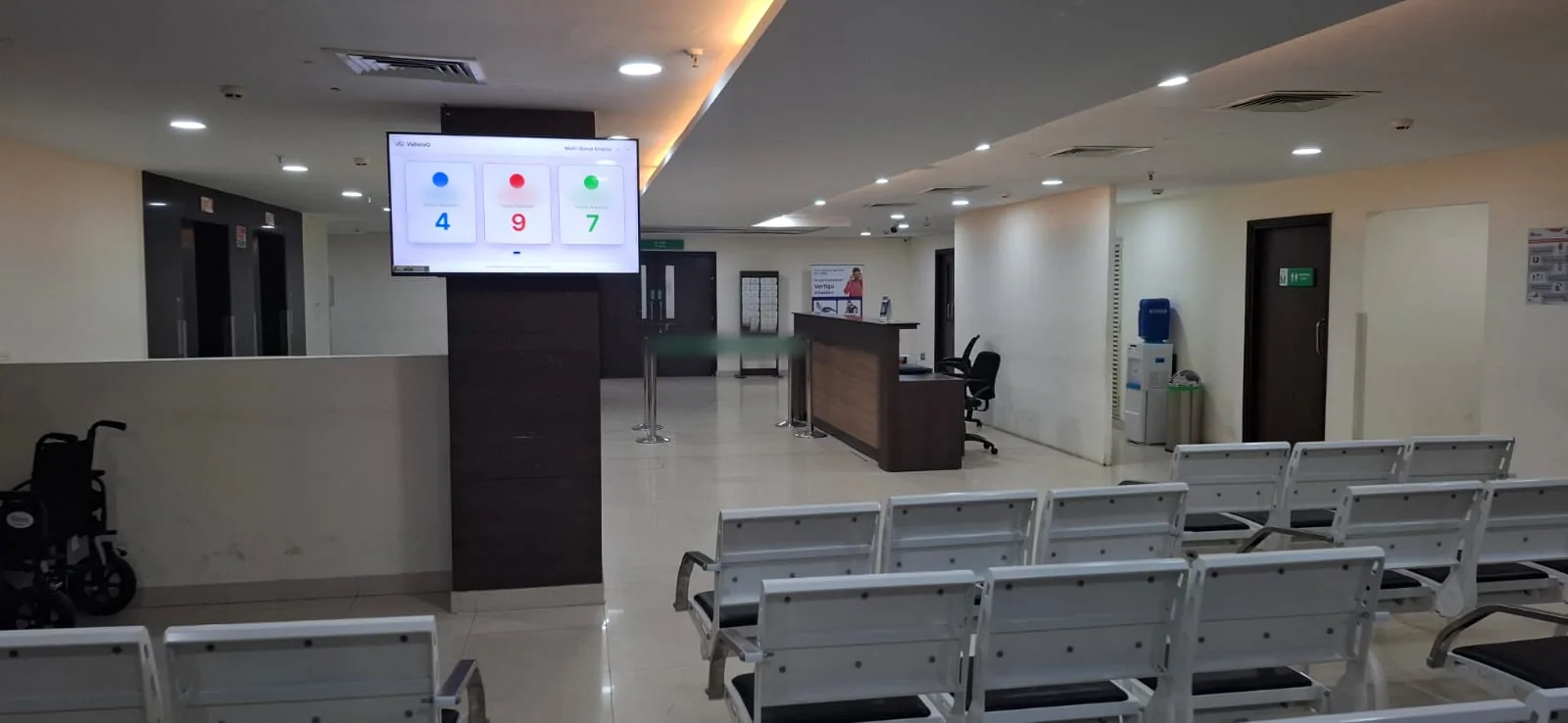 ValloraQ multi-queue display on a pillar in a hospital OPD waiting hall, showing tokens 4, 9 and 7 above rows of seats and the reception desk